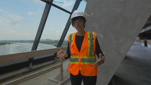 On voit Mme Mandalian, en tenue de travail, qui parle à la caméra. En arrière-plan, on voit les nouvelles fenêtres de la tour du stade et la structure en béton. On voit Mme Mandalian, en tenue de travail, qui parle à la caméra. En arrière-plan, on voit les nouvelles fenêtres de la tour du stade et la structure en béton.