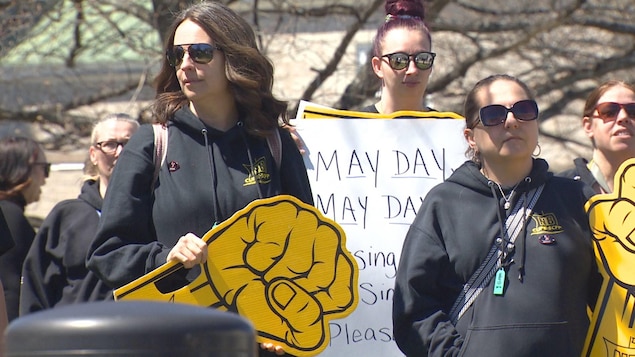Deux femmes en chandail à capuchon noir tiennent des poings fermés jaunes en carton. Une autre femme derrière tient une pancarte sur laquelle les mots « May Day May Day » sont écrits.