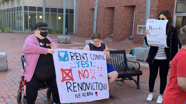 Une personne en fauteuil roulant, sur une place publique, tient une pancarte dont le message revendique un contrôle des loyers et la fin des rénovictions.