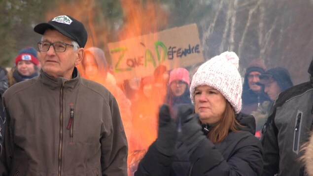 Un groupe de manifestants autour d'un feu en hiver. Une affiche « Zone à défendre » émerge en arrière. À l'avant, un homme regarde au loin et une femme applaudit.