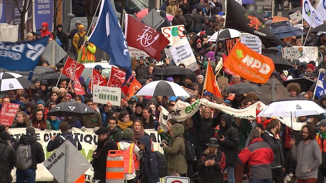 Plusieurs personnes brandissant des pancartes, des drapeaux ou des parapluies marchent dans une rue du centre-ville de Montréal.