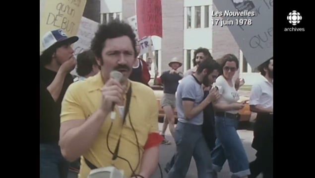 Un homme dans la manifestation tient un micro. À l'arrière, d'autres manifestants marchent.