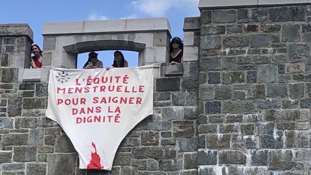 Des manifestantes tiennent une banderole au sommet des remparts sur la place d'Youville. On peut y lire « L'équité menstruelle pour saigner dans le dignité ».