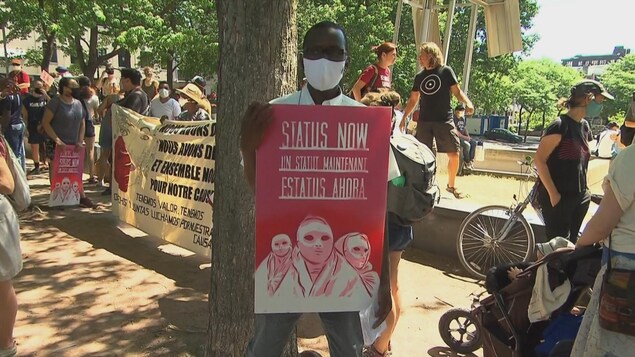 Un homme tient une affiche au milieu d'un rassemblement. on peut lire : « un statut maintenant » en plusieurs langues.