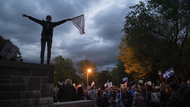 Une personne est debout sur un muret et agite un drapeau devant une foule.