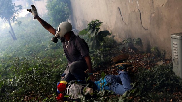 Un opposant au président Maduro vêtu d'un masque à gaz tente de venir en aide à un autre manifestant, blessé. Un opposant au président Maduro vêtu d'un masque à gaz tente de venir en aide à un autre manifestant, blessé.