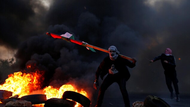 Deux hommes masqués de keffiehs se tiennent près de pneus en feu. L'un d'eux tient un drapeau palestinien, l'autre, lance des pierres avec un lance-pierre. Deux hommes masqués de keffiehs se tiennent près de pneus en feu. L'un d'eux tient un drapeau palestinien, l'autre, lance des pierres avec un lance-pierre.