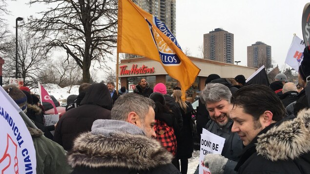 Des personnes rassemblées devant un Tim Hortons. Une femme fait un discours. Des personnes rassemblées devant un Tim Hortons. Une femme fait un discours.