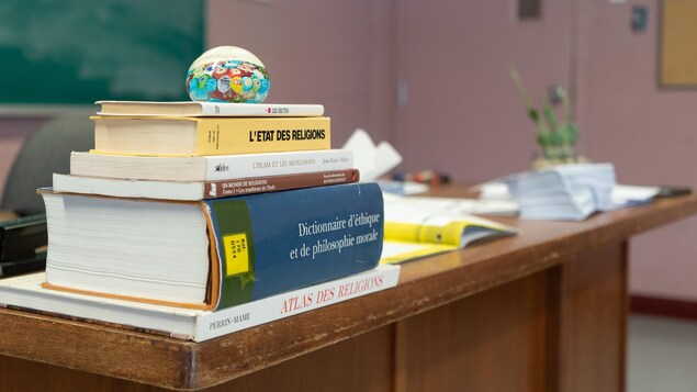 Une pile de manuels scolaires, un dictionnaire et un atlas sur un bureau en bois dans une salle de classe.
