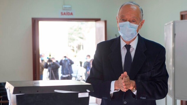Marcelo Rebelo de Sousa porte un masque chirurgical et joint les deux mains devant lui. À côté de lui se trouve une boîte de vote dans un bureau de scrutin.