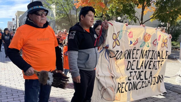 Jean-Paul Fontaine marchant aux côtés d'une banderole et arborant un chandail orange sur lequel il est écrit le mot survivant.