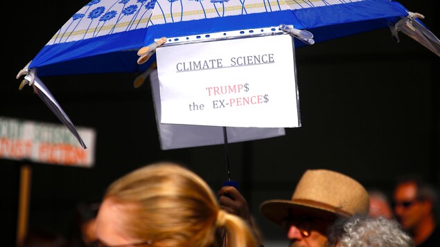 Un homme porte un parapluie où l'on peut lire : CLIMATE SCIENCE, TRUMP$ the EX-PENCE$ Un homme porte un parapluie où l'on peut lire : CLIMATE SCIENCE, TRUMP$ the EX-PENCE$