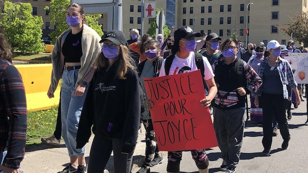 Une foule masquée pendant une manifestation. Une femme porte une pancarte où il est écrit «Justice pour Joyce».