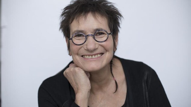 Portrait en couleur de l'autrice Marie Clark, sur fond pâle uni. Elle porte un chandail noir et des lunettes rondes, sourit et appuie son menton sur son poing droit.