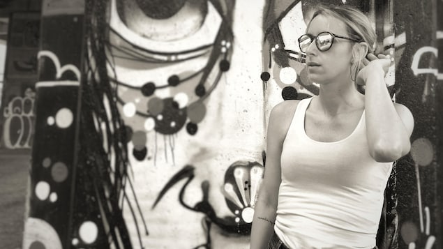 Portrait en noir et blanc de Marie St-Hilaire-Tremblay, de trois-quarts, en extérieur, devant une murale. Elle porte des lunettes de soleil miroir, une camisole blanche, et a la main gauche relevée derrière son oreille et a un tatouage sur l'intérieur de l'avant-bras droit.