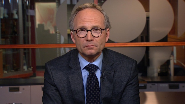 Un homme portant des lunettes ovales semble nous regarder droit dans les yeux. Il porte un veston-cravate et il a les cheveux grisonnants.