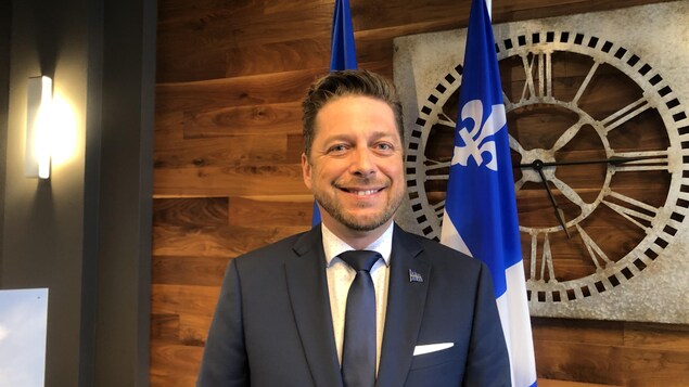 Martin Ouellet, devant un drapeau du Québec, sourit à la caméra.