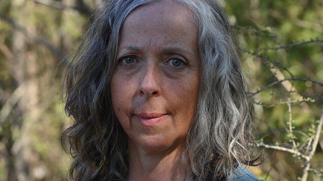 Portrait en couleur, en extérieur. La femme regarde la caméra. Elle a les cheveux gris, mi-longs, et lâchés.
