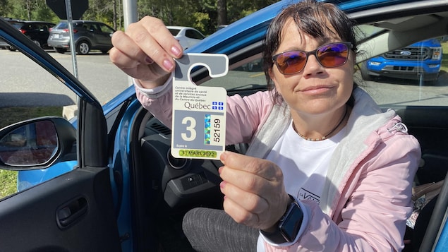 Martine Pombert tient dans ses mains une vignette de stationnement numérotée, assise dans son véhicule avec la portière côté conducteur entrouverte. 