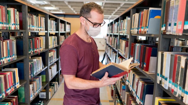 Un homme portant un masque feuillette un livre dans une bibliothèque.