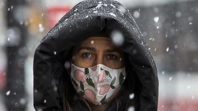 Une femme le visage couvert d'un masque et vêtue d'un manteau d'hiver à capuchon marche sous la neige à Toronto le 22 novembre 2020.