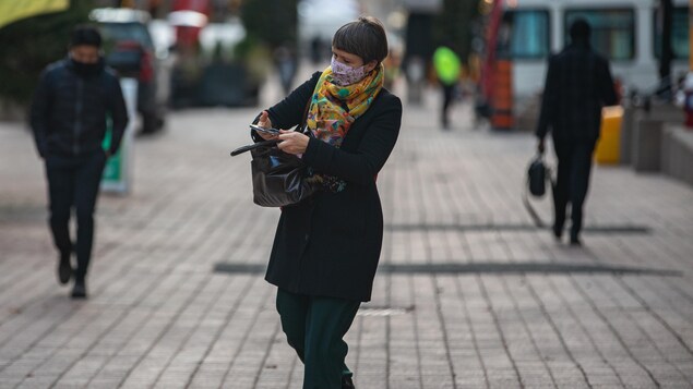 Une femme masquée marche en consultant son téléphone cellulaire.
