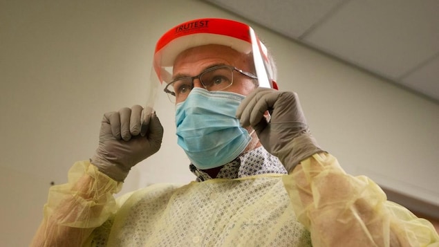 Un médecin porte l'équipement de protection individuelle : un masque, une blouse, des gants et une visière. 