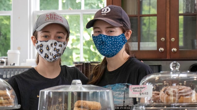 Deux jeunes femmes coiffées de casquette et le visage couvert d'un masque de tissu sont debout derrière le comptoir du café où elles travaillent.