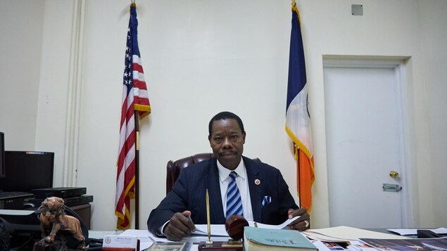 On voit M. Eugene de face, assis à son bureau. Des livres et des documents sont disposés devant lui. Derrière lui, une porte et le drapeau américain ainsi que celui de la Ville de New York. On voit M. Eugene de face, assis à son bureau. Des livres et des documents sont disposés devant lui. Derrière lui, une porte et le drapeau américain ainsi que celui de la Ville de New York.