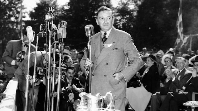 Maurice Duplessis prononce un discours debout derrière une série de micros sur pied lors de l'inauguration du pont Duplessis le 6 juin 1948. Derrière lui, on voit des photographes et le public, assis, venu assister à l'événement.