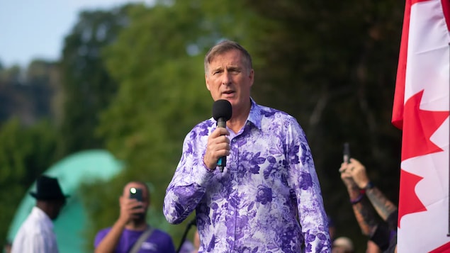 May hawak na mikropono si Maxime Bernier katabi ng watawat ng Canada.