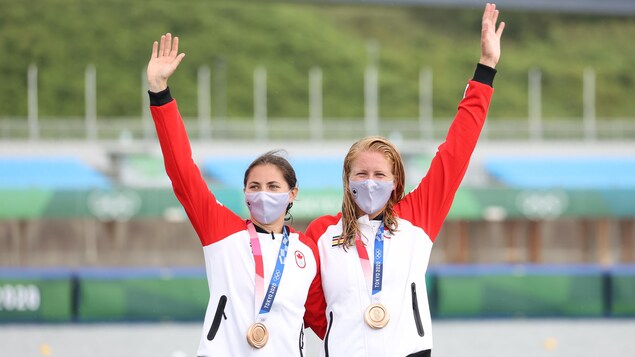 Deux femmes canadiennes masquées avec une médaille de bronze au cou se tiennent par la taille en saluant d'une main.