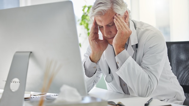 Un médecin assis devant son ordinateur regarde son bureau et se place les deux mains sur les tempes.