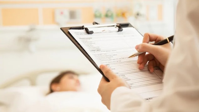 Un médecin écrit des notes dans le dossier d'un patient qu'on voit dans un lit d'hôpital en arrière-plan.