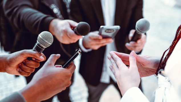 Des reporters tendent des micros et téléphone vers une femme qui parle. 