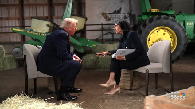 Un homme est assis au bout de sa chaise en gesticulant, une femme parle avec lui. Des tracteurs et des bottes de foin sont disposés en arrière-plan.