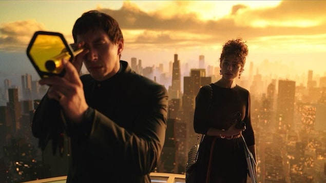 Un homme regarde dans une longue vue sur le toit d'un immeuble, avec une femme en arrière-plan.