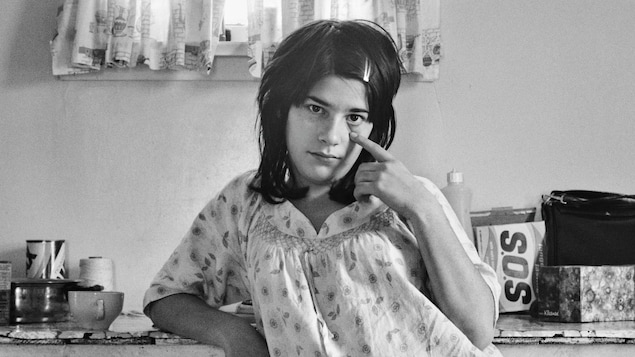 Photo en noir et blanc prise dans les années 1970 d'une jeune femme en jaquette, pieds nus, accoudée à un comptoir de cuisine couvert d'objets comme une boîte de céréales, une boîte de savon SOS et une radio. La femme étire la paupière inférieure de son oeil gauche avec son index.