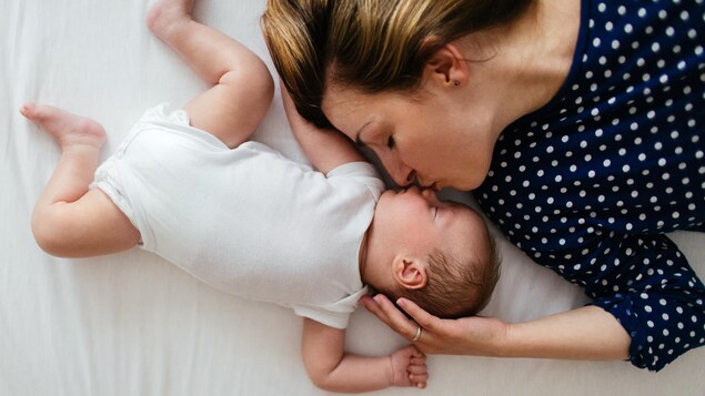 Une photo d'une mère en train d'embrasser son bébé endormi sur un grand lit.