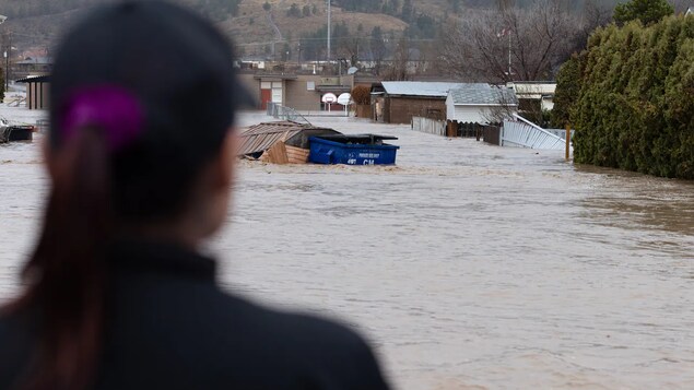 Une femme est debout devant un secteur inondé de Merritt qui comprend quelques maisons et des clôtures renversées.