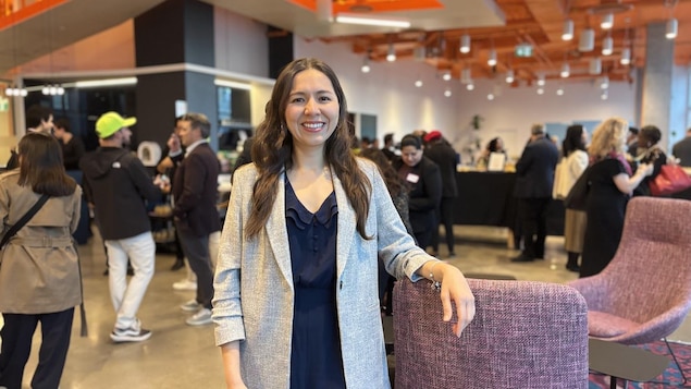 Esp: Mujer sonriente con chaqueta gris y vestido oscuro, de pie en un evento social interior.

Fr: Femme souriante en veste grise et robe foncée, posant lors d'un événement intérieur.