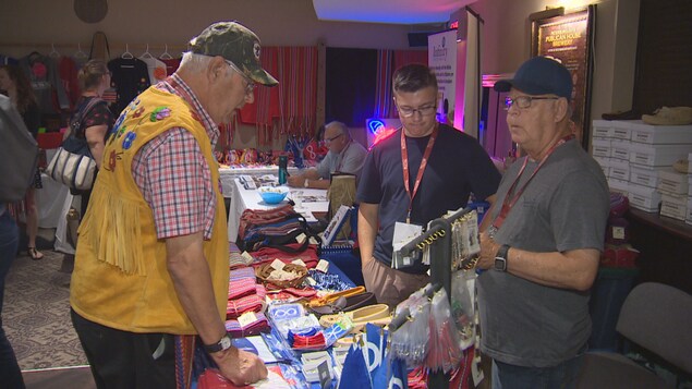 Des gens se promènent dans une foire artisane, avec des drapeaux métis et des ceintures fléchées sur une table. Des gens se promènent dans une foire artisane, avec des drapeaux métis et des ceintures fléchées sur une table.