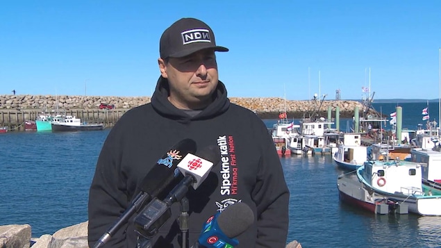 Mike Sack sur le quai, dos à une flotte de bateaux, devant plusieurs micros de différents médias.