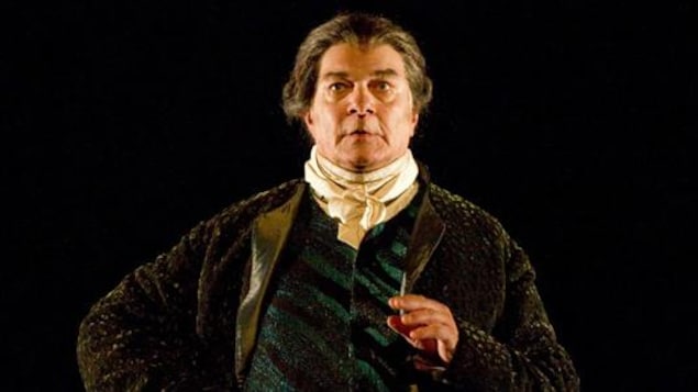 Un homme portant une perruque et un costume du 18e siècle s'exprime sur une scène de théâtre.