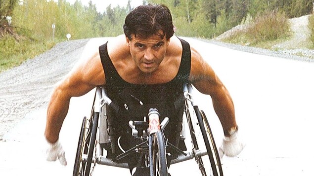 Michel Juteau roule sur une route à l'aide d'un fauteuil adapté à trois roues.