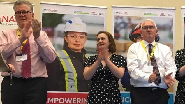 Une femme entourée de deux hommes à cravate applaudissant devant des affiches publicitaires