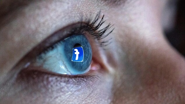 Une femme se tient devant un écran d'ordinateur alors que le logo du réseau social Facebook se réflète dans son œil.