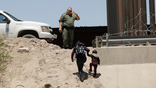 Une femme tient un enfant par la main et monte une pente en sable sur le sommet de laquelle se trouve l'agent frontalier.
