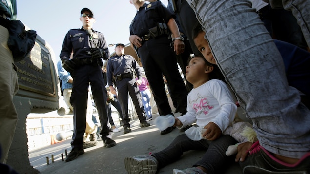 Deux jeunes enfants sont assis au sol, pendant que des garde-frontières américains sont debout parmi un groupe de migrants. Deux jeunes enfants sont assis au sol, pendant que des garde-frontières américains sont debout parmi un groupe de migrants.
