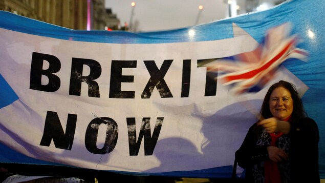 Une femme avec un drapeau du Royaume-Uni, derrière elle, une banderole indiquant "Brexit now"